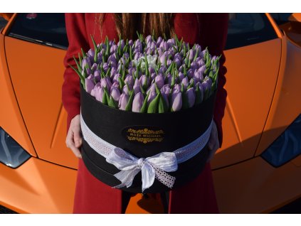 Luxusní box s fialovými tulipány, ozdobený bílou stuhou, umístěný na kapotě oranžového sportovního auta.