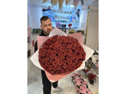 Muž drží třpytivý speciální puget ze 101 rudých růží, zabalený v elegantním růžovém papíru, v moderním interiéru květinářství.