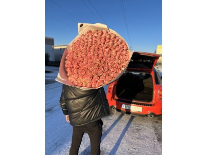 Muž v černé zimní bundě drží ohromující puget růžových růží "Love Pink" na pozadí modré oblohy a zaparkovaného červeného auta.
