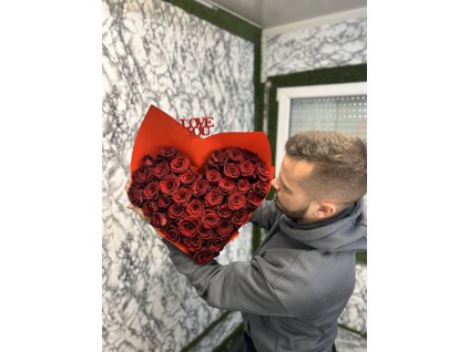 Muž v šedé mikině drží červený Valentýnský puget ve tvaru srdce, aranžovaný z rudých růží, na pozadí bílé zdobené stěny.