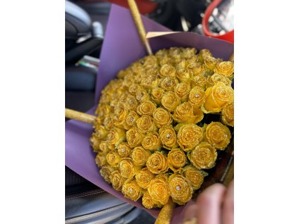 Třpytivý puget ze 101 žlutých růží, elegantně zabalený v fialovém papíru, prezentovaný venku s pozadím automobilu.