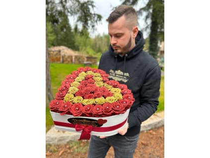 Muž drží třpytivý box ve tvaru srdce z rudých a žlutých růží, ozdobený elegantní červenou stuhou, na venkovním pozadí.