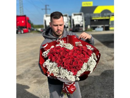 Kytice růží ve tvaru srdce zdobená gypsophilou, držená floristem na otevřeném prostranství před parkovištěm.