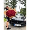 Velkolepý puget 201 červených růží v přírodním balení, držený mužem před černým luxusním vozem.