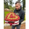 Muž drží třpytivý box ve tvaru srdce z rudých a žlutých růží, ozdobený elegantní červenou stuhou, na venkovním pozadí.