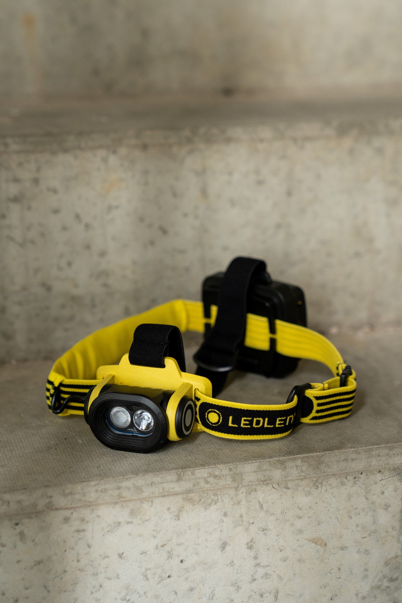 Ledlenser EXH6R žltá na betónovom schode — dve LED za oválnym sklom, žltý držiak, popruh Ledlenser a batériový box vzadu