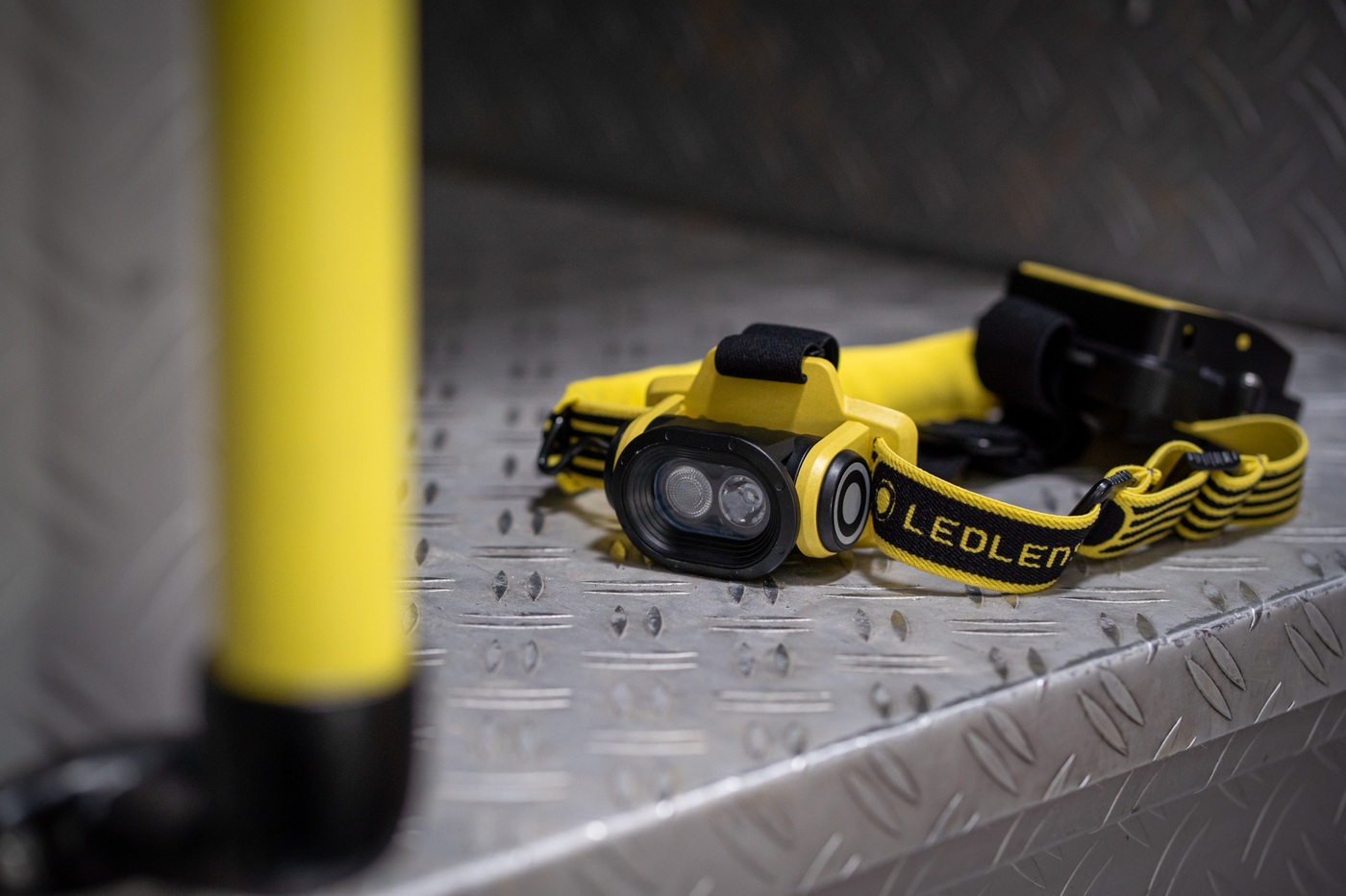 Ledlenser EXH6R žltá položená na ryhovanom hliníkovom plechu — hlava s dvoma LED vpredu, batériový box vzadu, žltá tyč vľavo