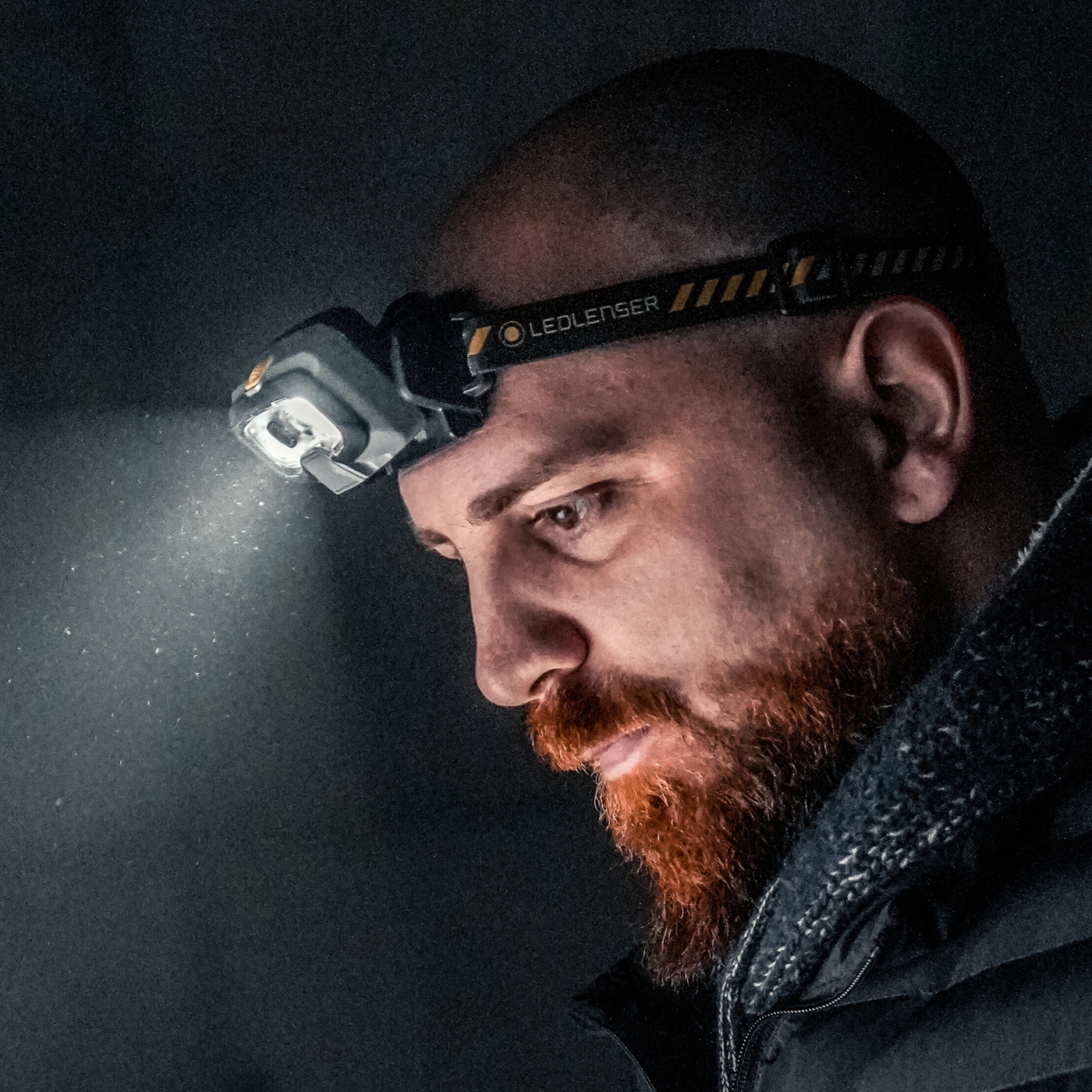 Ledlenser HF8R Work žltá na čele bradatého muža — čelovka svieti do prachu, popruh s nápisom Ledlenser a žltými pruhmi