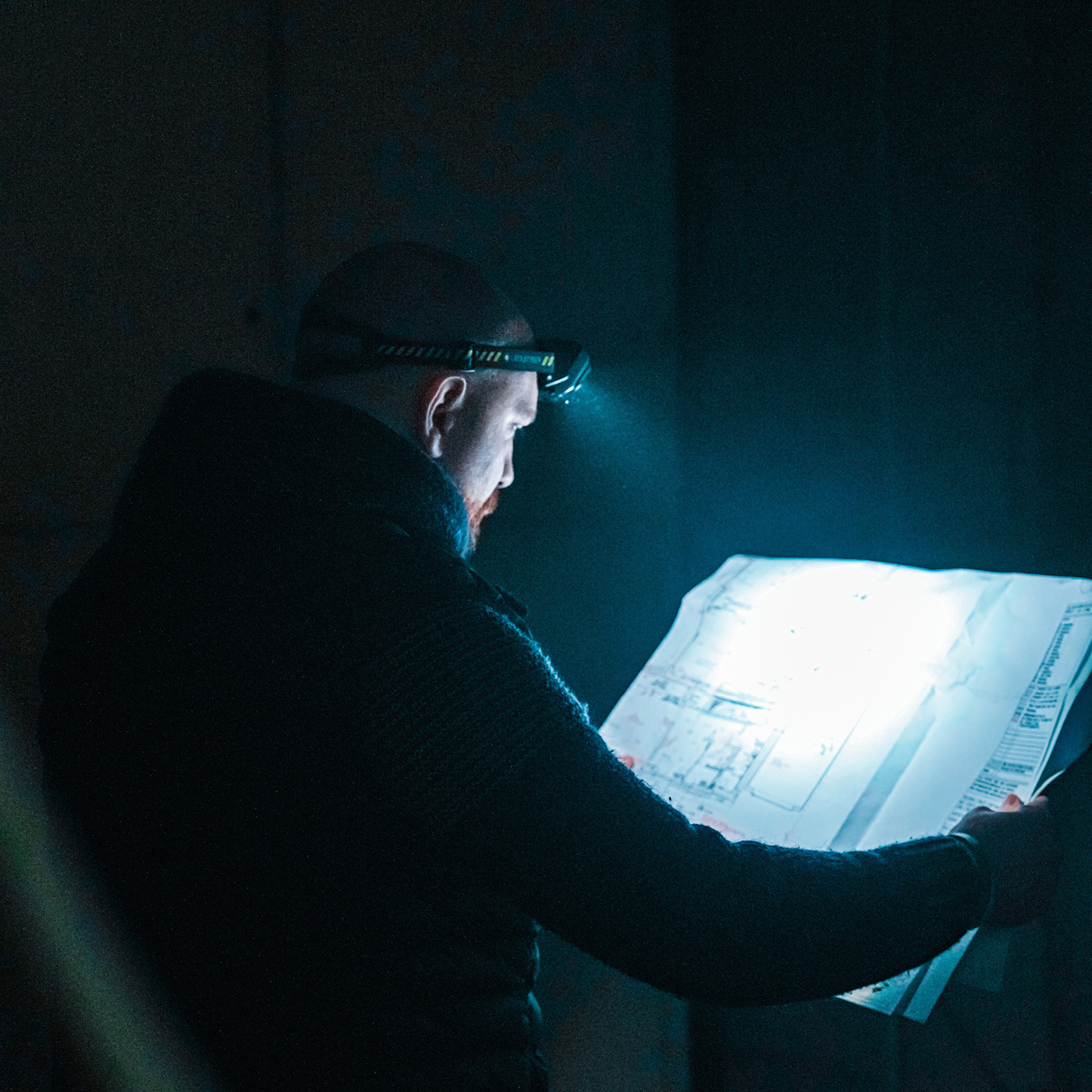 Ledlenser HF8R Work na hlave muža, ktorý si v tme číta stavebný výkres — kužeľ čelovky osvetľuje papier, popruh s pruhmi