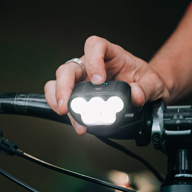 Ruka na riadidlách drží rozsvietené Magicshine MONTEER 5000S — päť LED svieti, telo sa zmestí do dlane