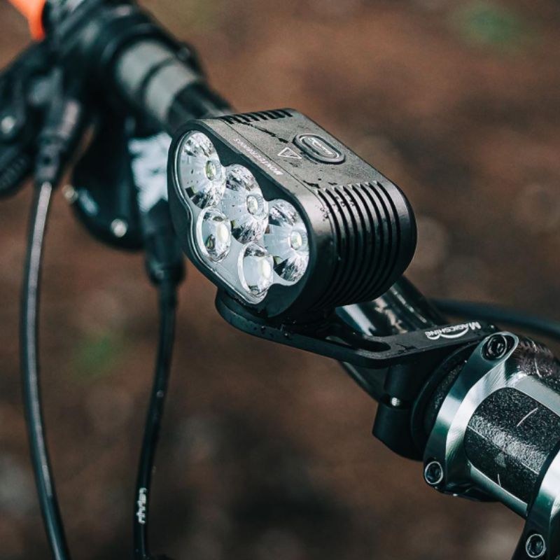 Magicshine MONTEER 8000S V2.0 upnuté na riadidlách horského bicykla v jesennom lese, vidno päť šošoviek a kábel k batérii
