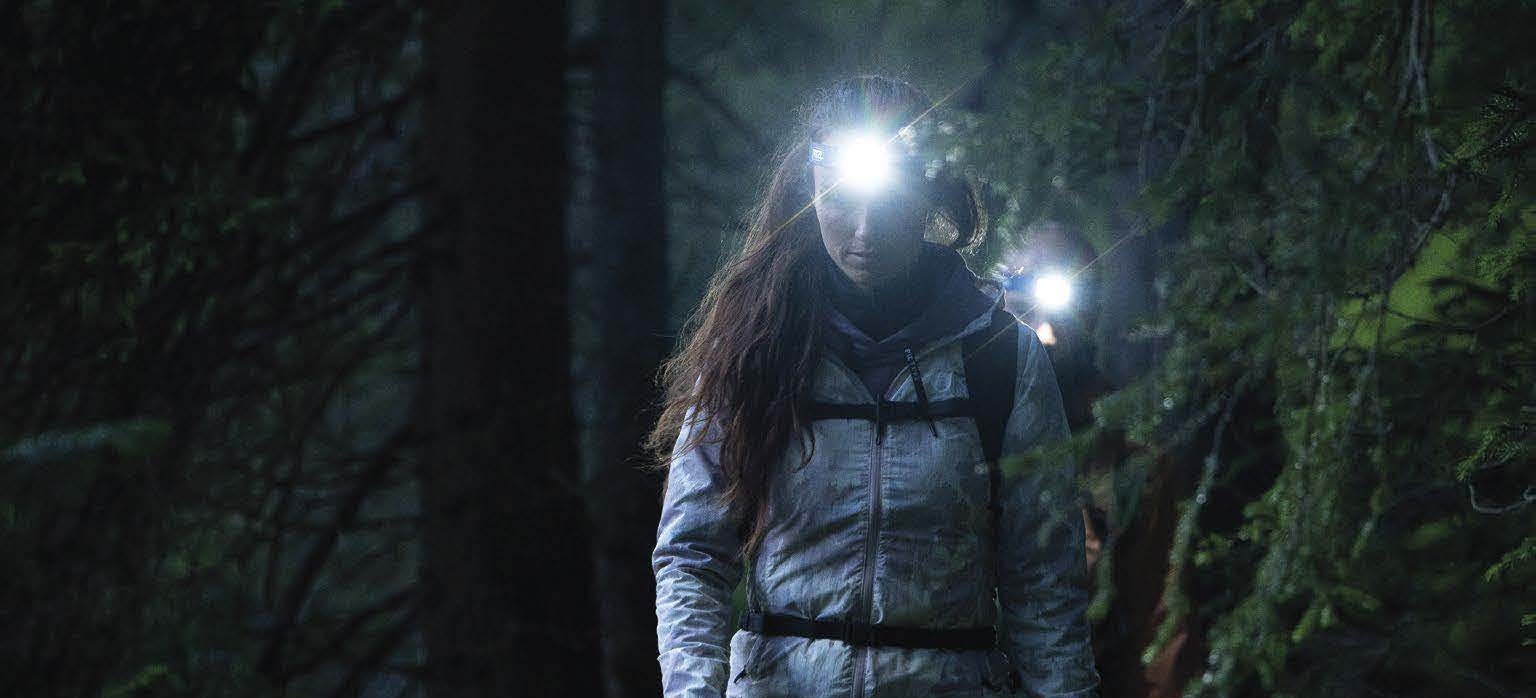 Turistka kráča v noci lesným chodníkom, čelovka Petzl jej osvetľuje cestu, za ňou ide druhý človek s rozsvietenou čelovkou