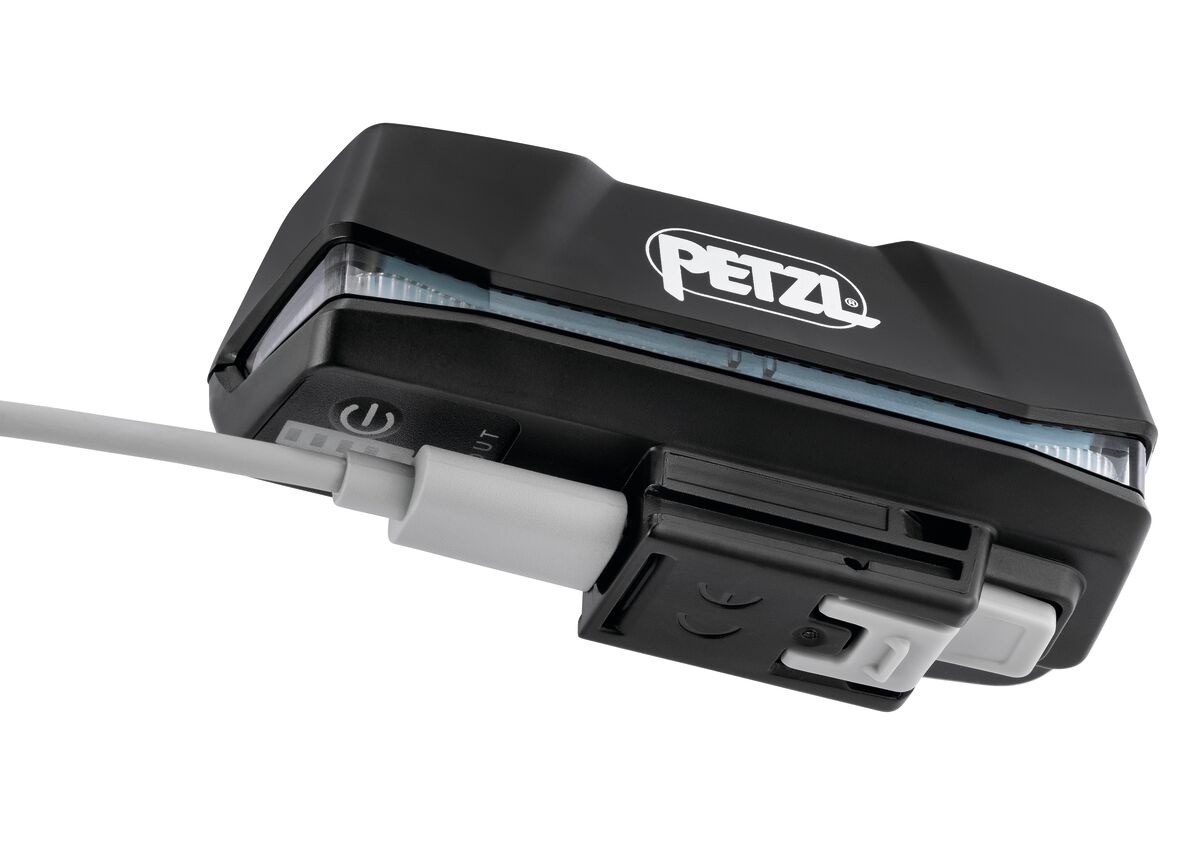 Akumulátor Petzl R1 zblízka s pripojeným sivým nabíjacím káblom, vedľa portu je tlačidlo a rad piatich značiek indikátora nabitia