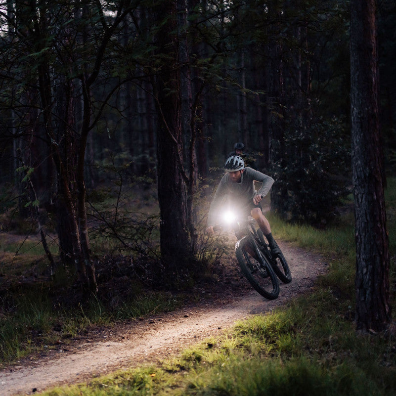 Cyklista na lesnom chodníku za šera so zapnutým predným svetlom Magicshine RAY 1600B na riadidlách