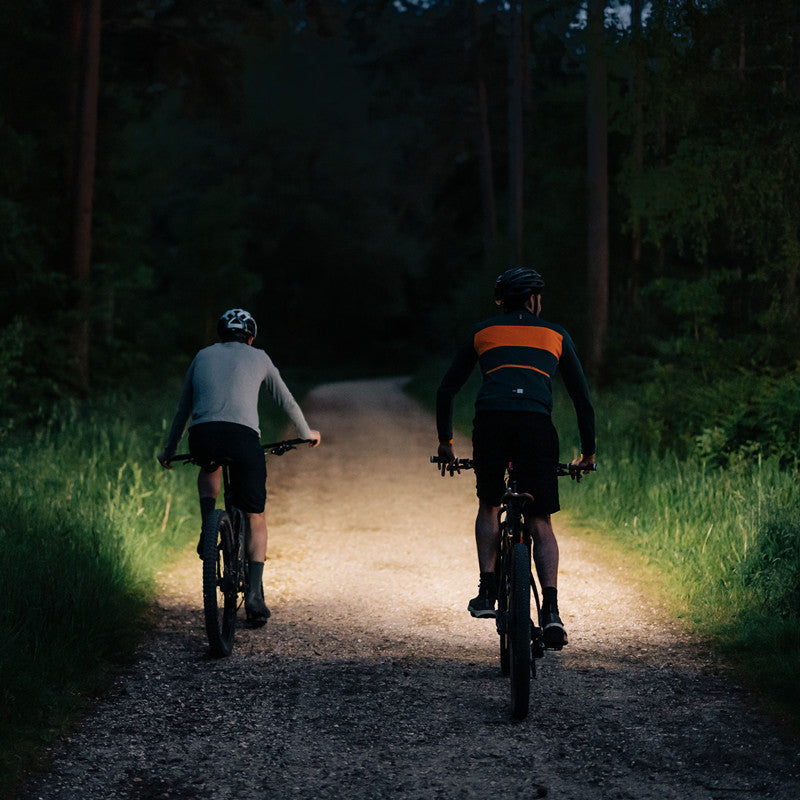 Dvaja cyklisti na lesnej šotolinovej ceste za šera osvetlenej predným cyklosvetlom Magicshine RAY 2600B