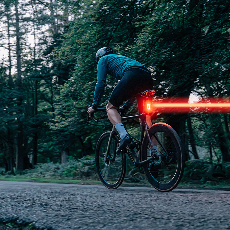 Fotografia výrobcu Magicshine: cyklista v lese za súmraku, zo zadného svetla SEEMEE 100AD vychádza výrazný červený lúč do strany