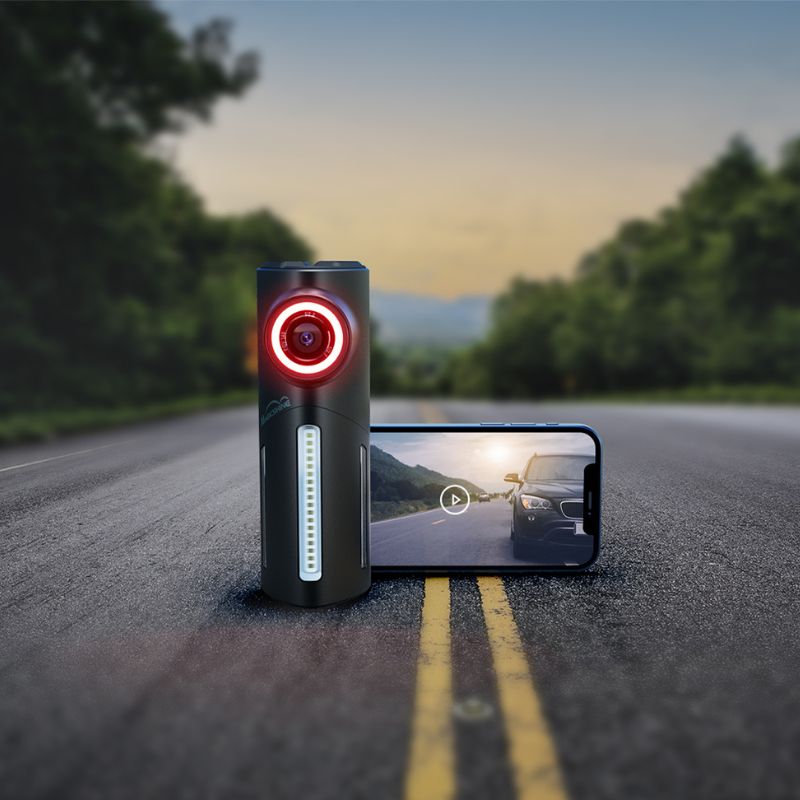 Magicshine SEEMEE DV stojí na ceste vedľa telefónu, na displeji je prehrávaný záznam auta idúceho za cyklistom