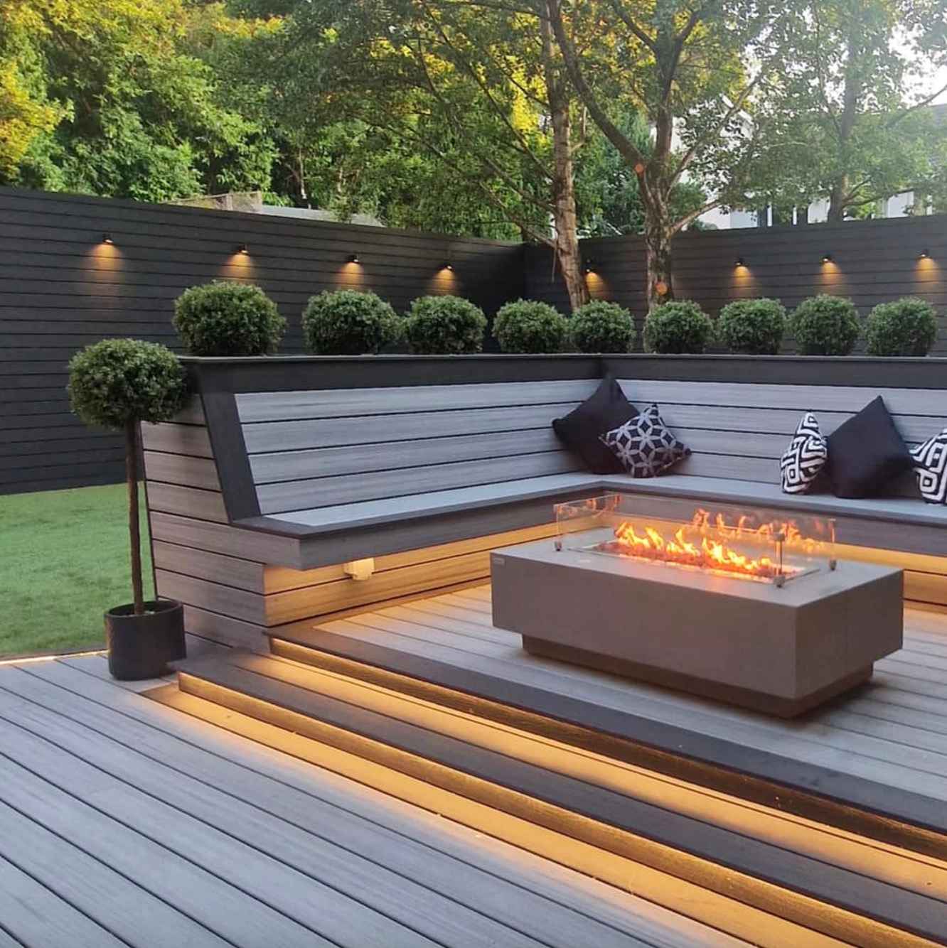 vstavane-sedenie-na-terase-z-svetlosivych-wpc-dosiek-s-led-osvetlenim-a-modernym-ohniskom-v-pozadi-sukromna-zahrada-so-zelenym-travnikom-pokojna-atmosfera