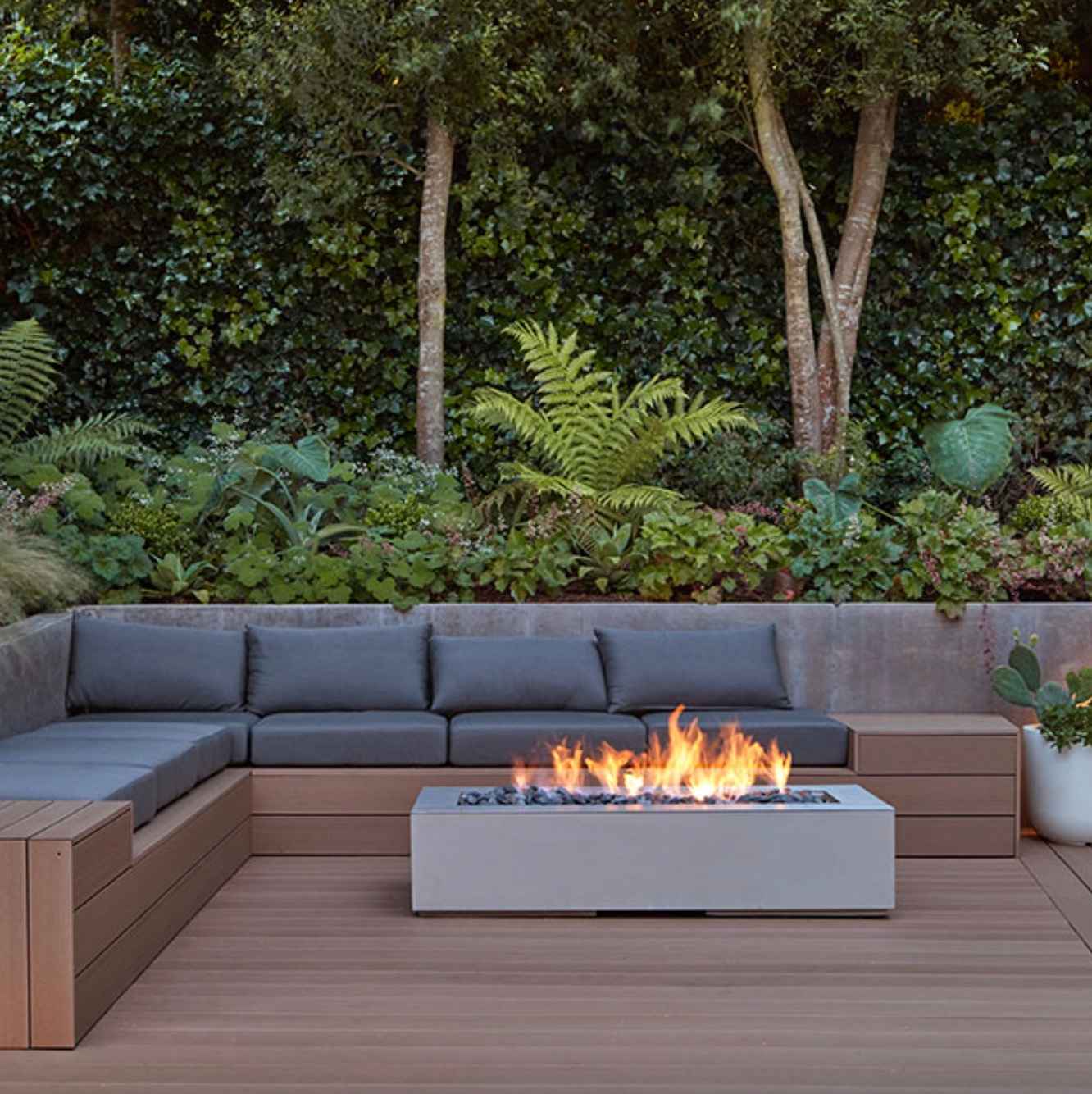nizky-moderny-zahradny-krb-na-terase-s-hnedych-wpc-dosiek-obklopeny-sivym-vstavanym-sedenim-a-hustou-zelenou-zahradou-v-pozadi-pokojne-vecerne-posedenie