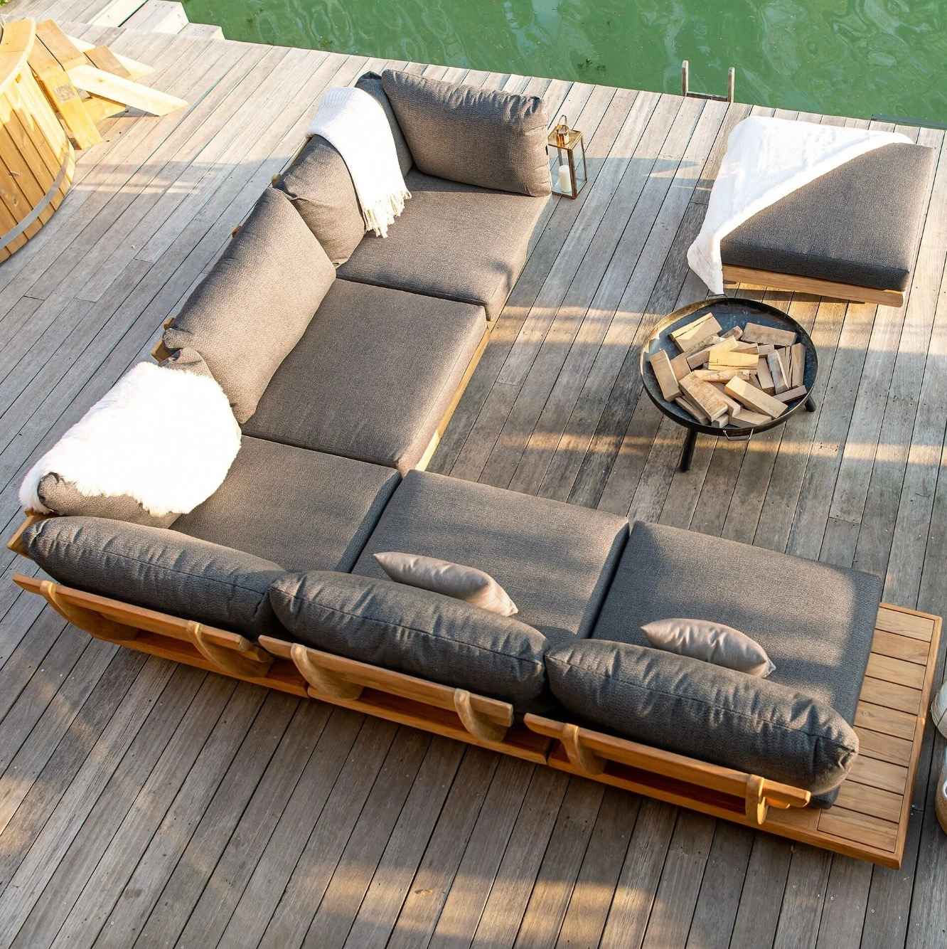zahradny-lounge-kutik-s-velkou-sedackou-v-tvare-l-na-drevenej-terase-pri-vode-s-kruhovym-ohniskom-a-dekoracnou-lampou-vecerna-pohoda-na-mole
