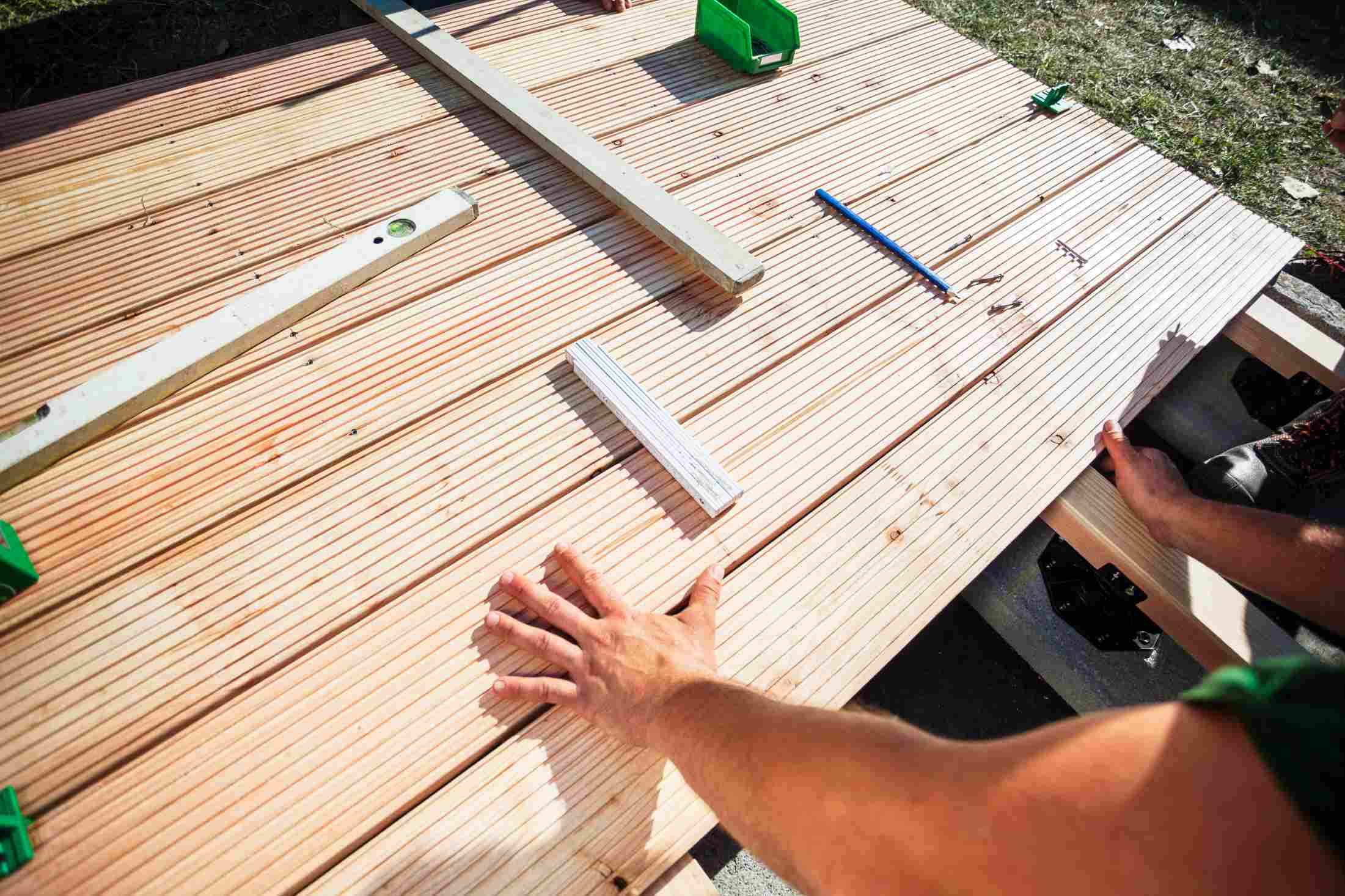 muzska-ruka-pridrziava-terasovu-dosku-pri-montazi-wpc-terasy-na-rosty-ulozene-na-viditelne-terce-okolo-je-naradie-vodovaha-a-spojovaci-material-na-povrchu-terasy
