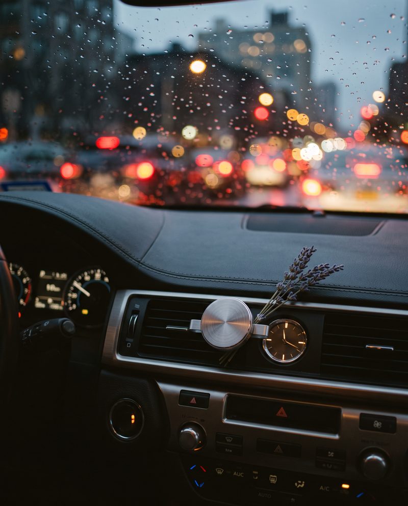 Autoinnenraum im abendlichen Stau mit einem Medaillon am Lüftungsgitter und einem Lavendelzweig, Regen auf der Scheibe
