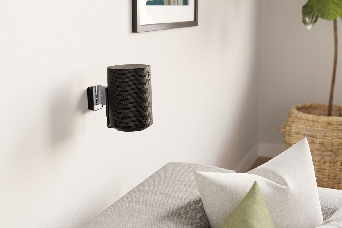 Reproduktor Sonos Era 100 v černé barvě zavěšený na světlé stěně obývacího pokoje na držáku Vogel's SWM 4111, vedle obraz a pohovka s polštáři