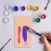 Bastelbrett wird mit bunter Farbe bemalt, Farbkleckse im Hintergrund